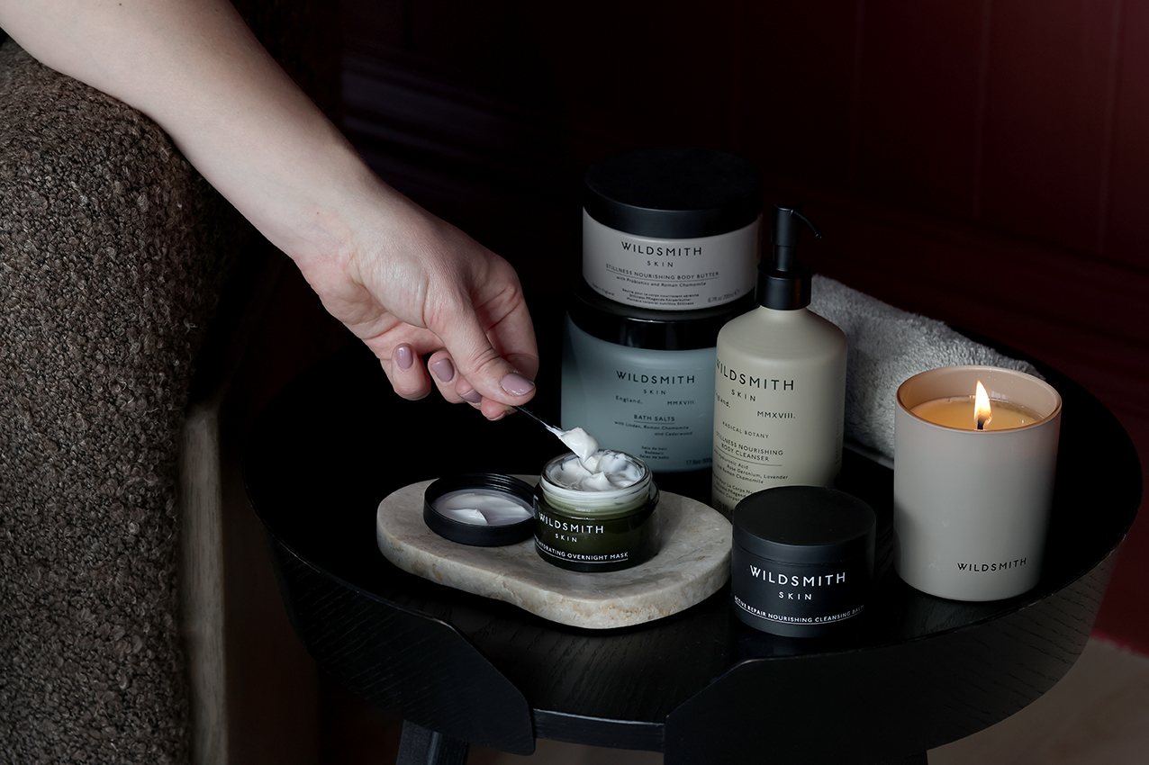Luxury Wildsmith lit candle and skin care products on a black bedside table, a ladies hand scooping the contents of face cream
